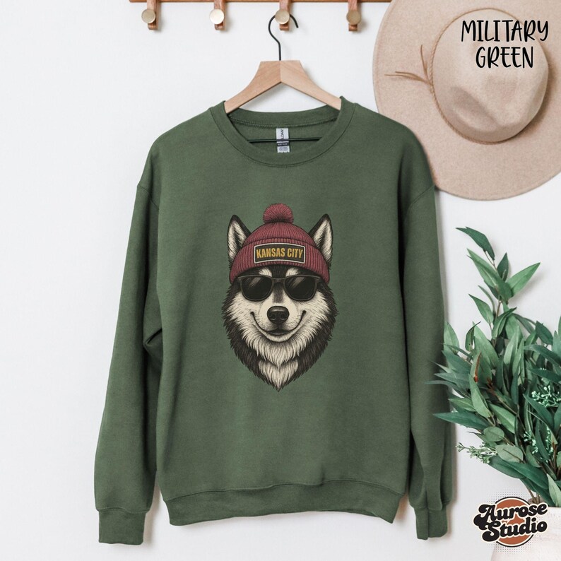Op de afbeelding: Militairgroen sweatshirt met een afbeelding van een husky met een zonnebril en een bordeauxrode muts met de tekst "Kansas City". Het sweatshirt hangt aan een houten hanger.