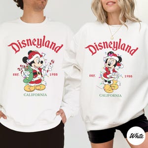 K&ouml;nnte beinhalten: Wei&szlig;e Sweatshirts mit einem Disneyland-Motiv mit Mickey und Minnie Mouse in Weihnachtskleidung. Die Sweatshirts haben den Text "Disneyland" in roter Schrift, "EST. 1955" und "CALIFORNIA" in Gr&uuml;n.