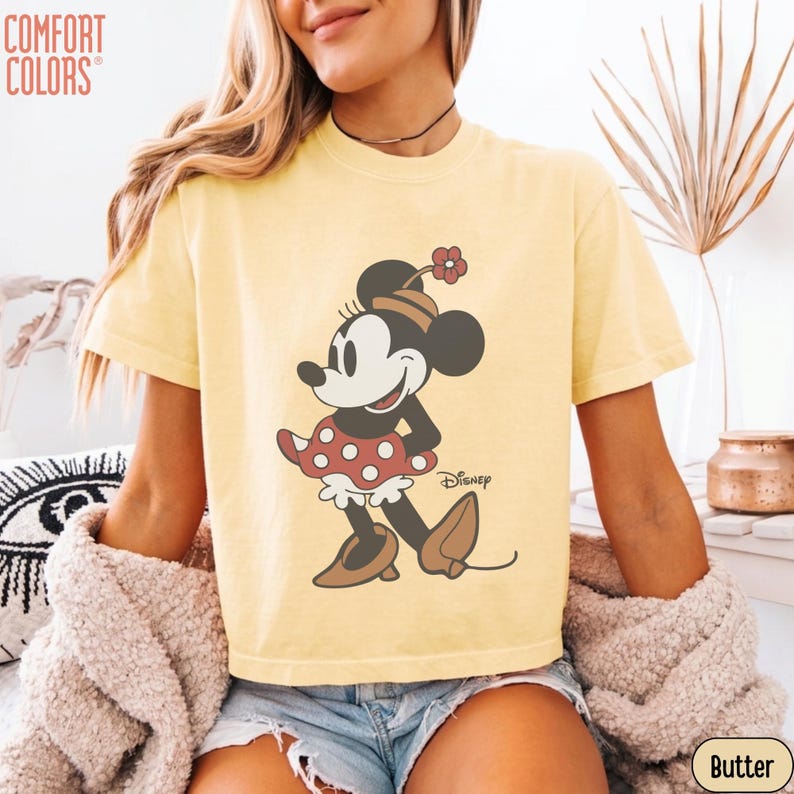 May include: Butter yellow Comfort Colors t-shirt featuring a Minnie Mouse graphic. The cartoon character is depicted in a red polka dot dress, brown shoes, and a matching hat with a flower. The Disney logo is printed on the shirt.