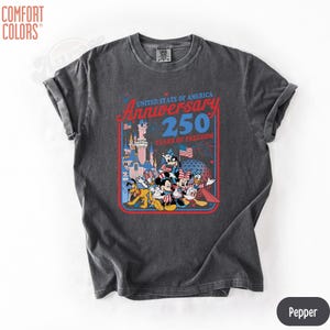 May include: Pepper-colored t-shirt featuring a graphic celebrating the United States of America's 250th anniversary. The design includes iconic Disney characters in patriotic attire, a castle, and the American flag. The shirt is from Comfort Colors.
