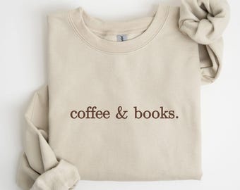May include: A beige sweatshirt with the text "coffee & books." in brown lettering. The sweatshirt has long sleeves and a crew neck. A matching scrunchie is placed on the right side of the sweatshirt.