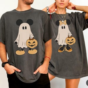May include: Two charcoal gray t-shirts featuring a graphic of a ghost wearing mouse ears and holding a jack-o'-lantern. One ghost has plain ears, while the other wears a polka-dot bow and orange shoes. The shirts have a relaxed, short-sleeve fit.