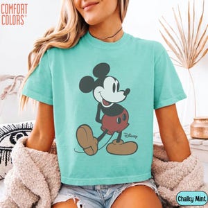 May include: A turquoise Comfort Colors t-shirt featuring a classic Mickey Mouse graphic. The cartoon character is depicted in black, red, and tan, with the Disney logo below. The shirt is labeled "Chalky Mint".