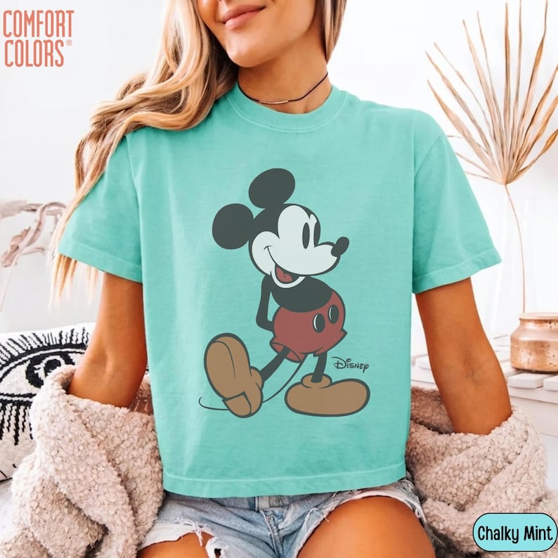 May include: A turquoise Comfort Colors t-shirt featuring a classic Mickey Mouse graphic. The cartoon character is depicted in black, red, and tan, with the Disney logo below. The shirt is labeled "Chalky Mint".