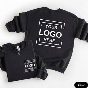 Pode incluir: Duas camisolas pretas com o texto "YOUR LOGO HERE" em branco. Uma camisola tem o texto na frente e a outra nas costas. As camisolas são exibidas num fundo branco.