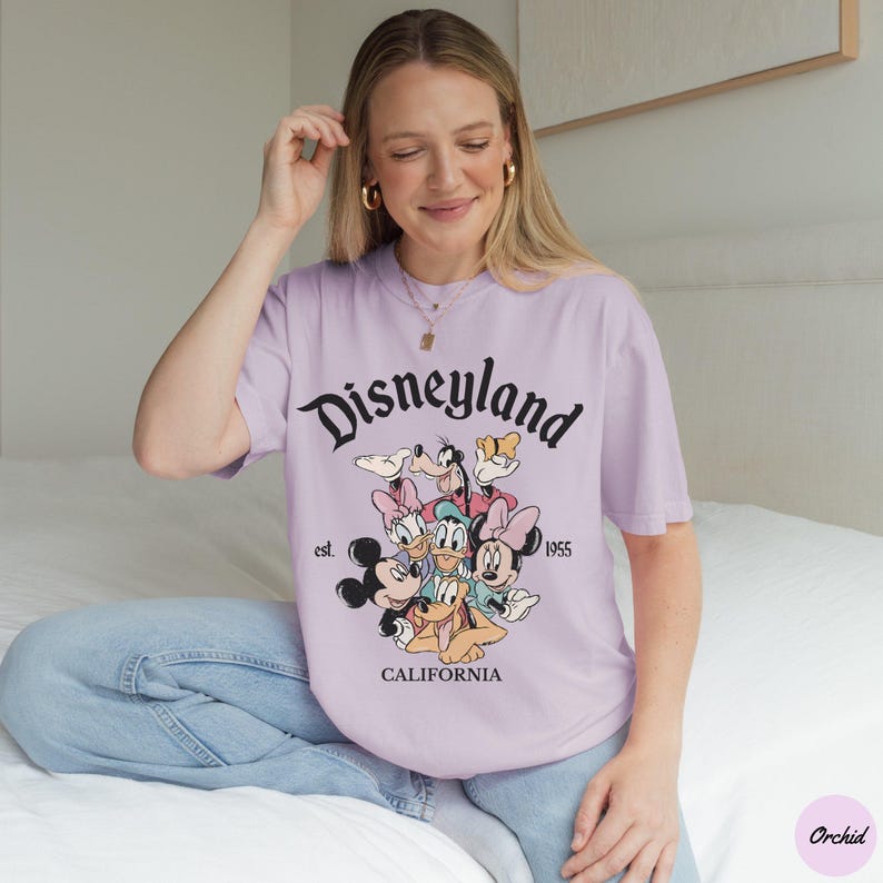 May include: A light purple t-shirt with the word "Disneyland" in black script, featuring a graphic of Disney characters. The shirt also includes the text "est. 1955" and "CALIFORNIA".