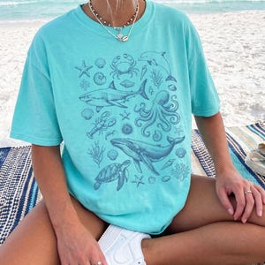 Pode incluir: Uma t-shirt de manga curta verde menta com uma impress&atilde;o gr&aacute;fica azul escura de v&aacute;rias criaturas marinhas, incluindo um tubar&atilde;o, polvo, baleia, tartaruga marinha, caranguejo, lagosta, estrelas-do-mar e conchas num design central.