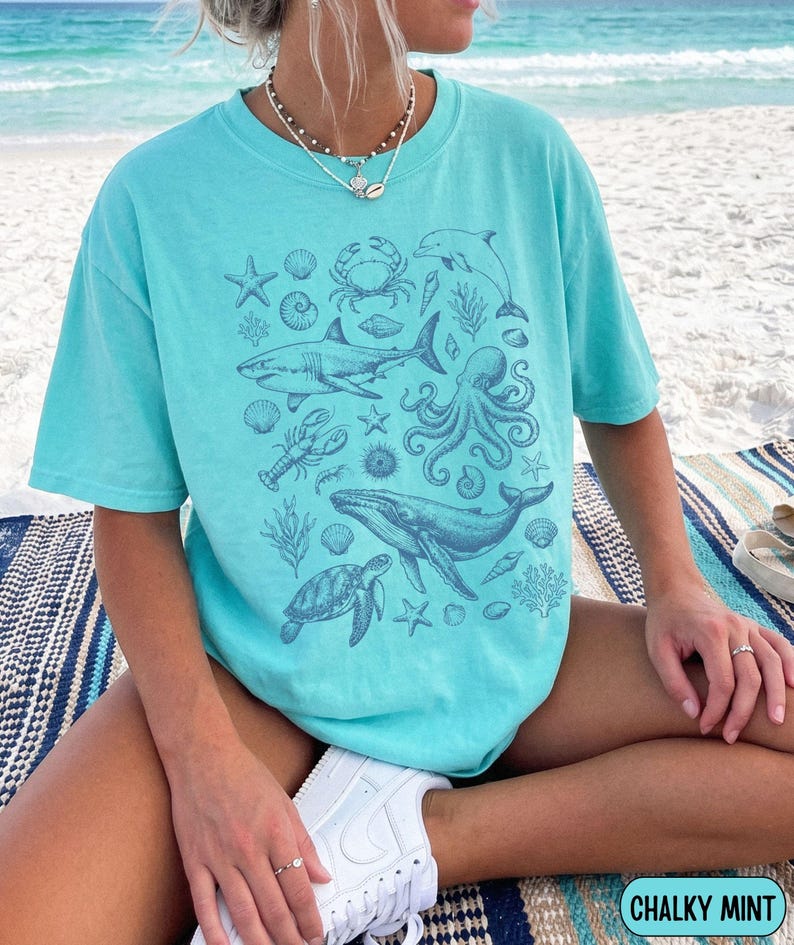 Pode incluir: Uma t-shirt de manga curta verde menta com uma impress&atilde;o gr&aacute;fica azul escura de v&aacute;rias criaturas marinhas, incluindo um tubar&atilde;o, polvo, baleia, tartaruga marinha, caranguejo, lagosta, estrelas-do-mar e conchas num design central.
