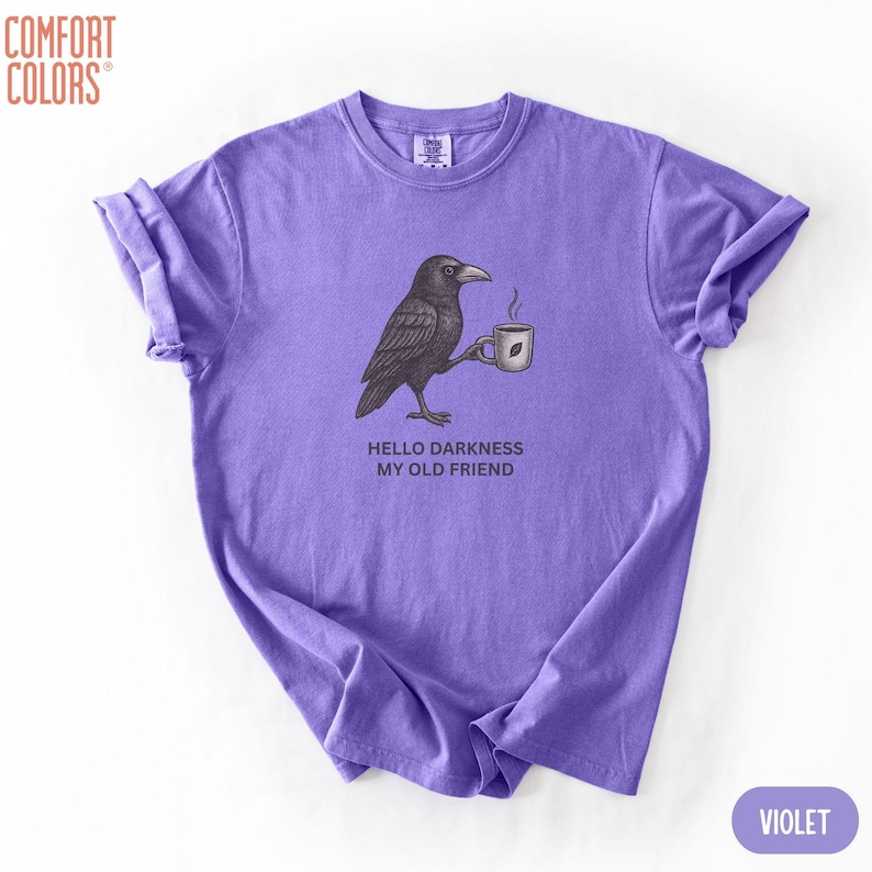 May include: A violet short-sleeve t-shirt featuring a black and white graphic of a crow holding a steaming coffee mug. Below the crow, the text reads, "HELLO DARKNESS MY OLD FRIEND." The shirt has a crew neckline and a Comfort Colors brand label.
