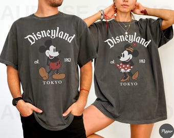 Puede incluir: Dos camisetas gris carbón con el logotipo de Disneyland y ilustraciones de Mickey y Minnie Mouse. Las camisetas incluyen el texto "Disneyland", "est. 1983" y "TOKYO". La camiseta de Mickey Mouse tiene un pantalón corto rojo, y la camiseta de Minnie Mouse tiene una falda de lunares rojos.