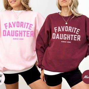 K&ouml;nnte beinhalten: Zwei Sweatshirts, eines in Hellrosa und eines in Weinrot, mit dem Text "FAVORITE DAUGHTER SINCE 1990" in einer verwaschenen Schriftart. Die Sweatshirts werden an Models mit schwarzen Shorts gezeigt.
