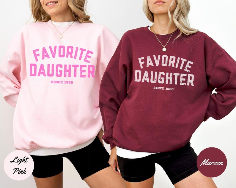 K&ouml;nnte beinhalten: Zwei Sweatshirts, eines in Hellrosa und eines in Weinrot, mit dem Text "FAVORITE DAUGHTER SINCE 1990" in einer verwaschenen Schriftart. Die Sweatshirts werden an Models mit schwarzen Shorts gezeigt.