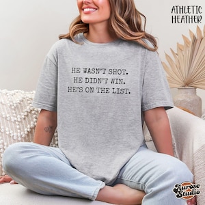 Może przedstawiać: Szara koszulka Athletic Heather z napisem "HE WASN'T SHOT. HE DIDN'T WIN. HE'S ON THE LIST." Tekst jest wydrukowany na czarno. Koszulka ma okrągły dekolt i luźny kr&oacute;j. Słowa "Athletic Heather" są wydrukowane w prawym g&oacute;rnym rogu.