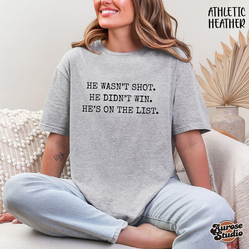 Może przedstawiać: Szara koszulka Athletic Heather z napisem "HE WASN'T SHOT. HE DIDN'T WIN. HE'S ON THE LIST." Tekst jest wydrukowany na czarno. Koszulka ma okrągły dekolt i luźny kr&oacute;j. Słowa "Athletic Heather" są wydrukowane w prawym g&oacute;rnym rogu.