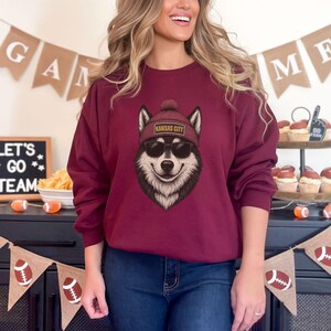 Op de afbeelding: Bordeaux sweatshirt met een afbeelding van een lachende wolf met een rode muts met de tekst "Kansas City" en een zonnebril. De sweatshirt heeft een ronde hals. De afbeelding is van een persoon die de sweatshirt draagt.