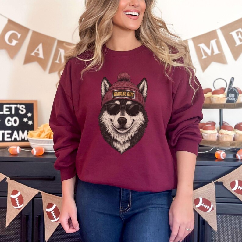 Op de afbeelding: Bordeaux sweatshirt met een afbeelding van een lachende wolf met een rode muts met de tekst "Kansas City" en een zonnebril. De sweatshirt heeft een ronde hals. De afbeelding is van een persoon die de sweatshirt draagt.