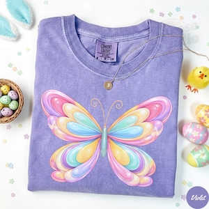 May include: A violet Comfort Colors t-shirt featuring a large, colorful butterfly design. The butterfly has pastel wings in shades of pink, yellow, blue, and green. A silver necklace with a pendant rests on the shirt. Easter eggs and a chick are in the background.