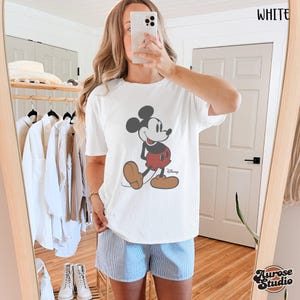 May include: White t-shirt featuring a classic Mickey Mouse graphic. The cartoon character is depicted in black, red, and brown. The word "Disney" is printed below the character. The word "WHITE" is printed in the upper right corner.