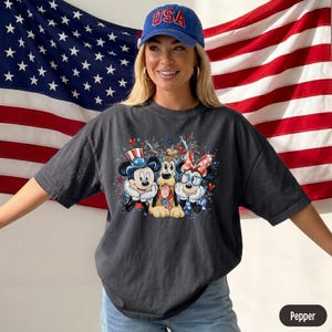May include: Dark gray t-shirt featuring Mickey Mouse, Pluto, and Minnie Mouse in patriotic attire. Mickey wears a top hat, Minnie a bow, and Pluto a red, white, and blue collar. The shirt is paired with a blue baseball cap with "USA" on it.