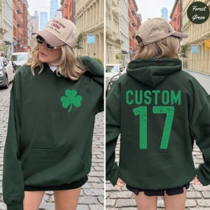 K&ouml;nnte beinhalten: Forstgr&uuml;ner Kapuzenpullover mit einer Vordertasche und einer gr&uuml;nen Kleeblatt-Grafik. Auf der R&uuml;ckseite des Hoodies steht das Wort "CUSTOM" &uuml;ber der Zahl "17" in Gr&uuml;n. Der Hoodie wird mit einer beigen Baseballkappe getragen.