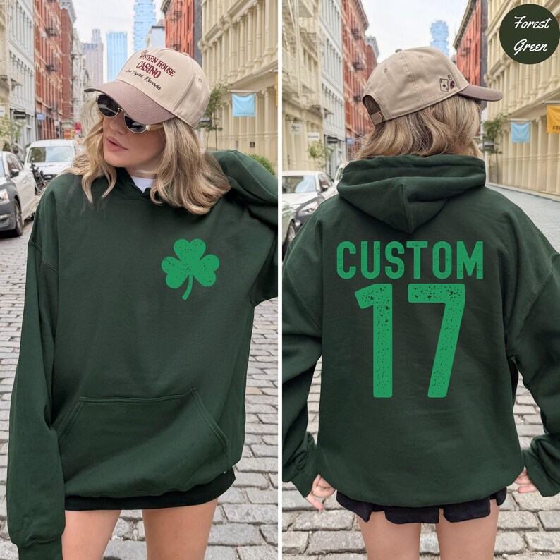 K&ouml;nnte beinhalten: Forstgr&uuml;ner Kapuzenpullover mit einer Vordertasche und einer gr&uuml;nen Kleeblatt-Grafik. Auf der R&uuml;ckseite des Hoodies steht das Wort "CUSTOM" &uuml;ber der Zahl "17" in Gr&uuml;n. Der Hoodie wird mit einer beigen Baseballkappe getragen.
