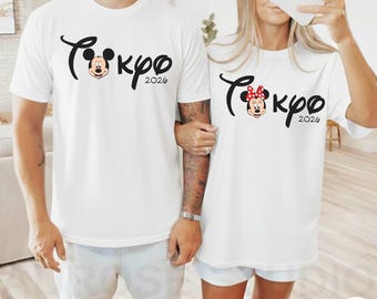 Puede incluir: Camisetas blancas con un diseño negro "Tokyo 2026" con Mickey y Minnie Mouse. El diseño de Mickey Mouse tiene un contorno negro con una cara sonriente. El diseño de Minnie Mouse tiene un lazo rojo. Las camisetas son usadas por dos personas.