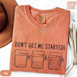 May include: Coral-colored t-shirt with the phrase "DON'T GET ME STARTED!" in black. Below the text are three jar illustrations with tags. A gold necklace with a pendant is draped on the shirt. The shirt has a Comfort Colors tag.