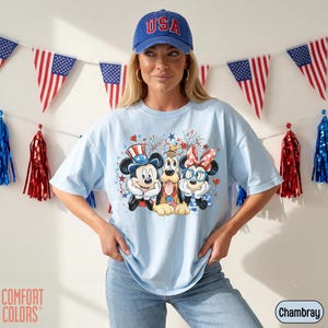 May include: Light blue t-shirt featuring Mickey Mouse, Pluto, and Minnie Mouse in patriotic attire. The shirt has a graphic print with fireworks and stars. A blue baseball cap with "USA" is worn. American flag bunting and red, white, and blue decorations are in the background.