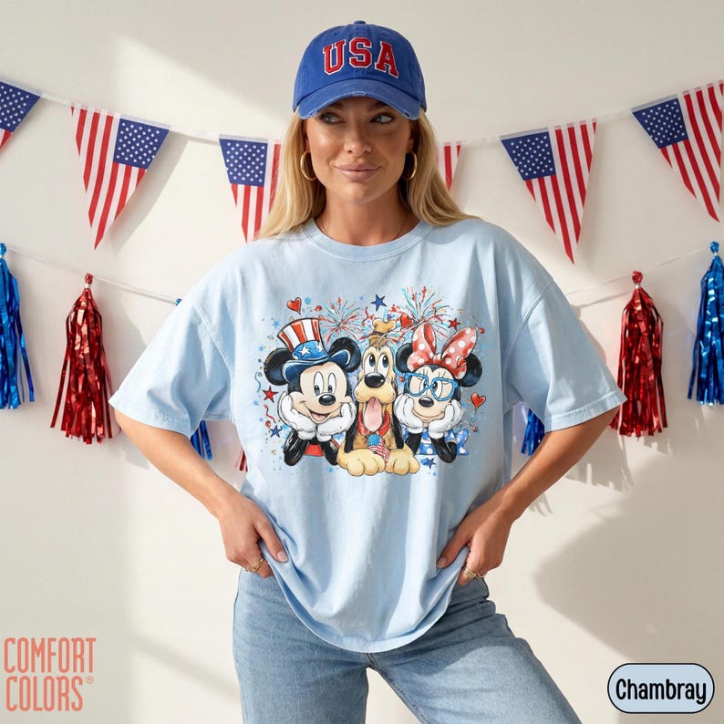 May include: Light blue t-shirt featuring Mickey Mouse, Pluto, and Minnie Mouse in patriotic attire. The shirt has a graphic print with fireworks and stars. A blue baseball cap with "USA" is worn. American flag bunting and red, white, and blue decorations are in the background.