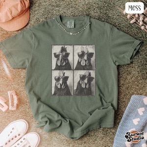 Op de afbeelding: Mosgroen t-shirt met een vierpaneelsafbeelding van kippen met zonnebrillen. Het shirt heeft een ronde hals en korte mouwen. Een roze corduroy pet en witte sneakers zijn zichtbaar in de afbeelding.