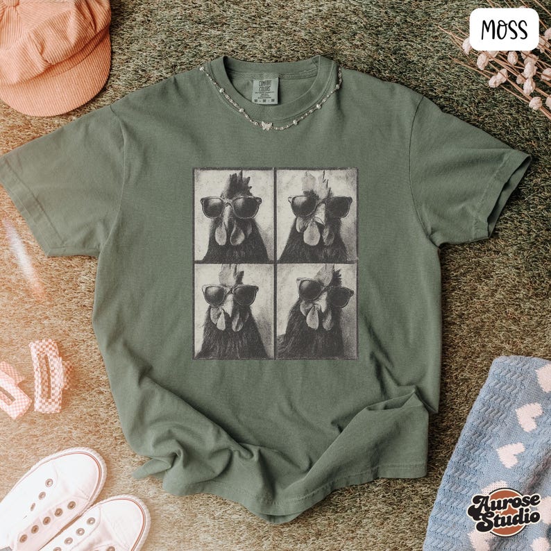 Op de afbeelding: Mosgroen t-shirt met een vierpaneelsafbeelding van kippen met zonnebrillen. Het shirt heeft een ronde hals en korte mouwen. Een roze corduroy pet en witte sneakers zijn zichtbaar in de afbeelding.