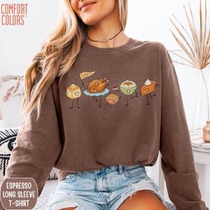 May include: A brown long-sleeve t-shirt featuring a graphic of cartoon Thanksgiving food items including a turkey holding a flag that says Thankful, a dinner roll, mashed potatoes, stuffing, and a slice of pumpkin pie with whipped cream.