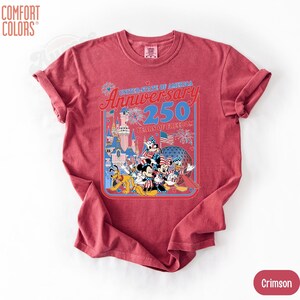 May include: Crimson-colored t-shirt featuring a graphic celebrating the 250th anniversary of the United States of America. The design includes cartoon characters, a castle, fireworks, and the text "United State of America Anniversary 250 Years of Freedom."