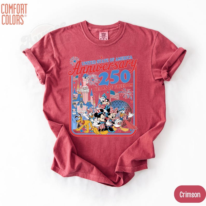 May include: Crimson-colored t-shirt featuring a graphic celebrating the 250th anniversary of the United States of America. The design includes cartoon characters, a castle, fireworks, and the text "United State of America Anniversary 250 Years of Freedom."