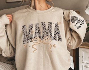 Può includere: Una felpa girocollo color sabbia con la scritta MAMA in stampa leopardata sul petto. Un fiocco beige è posizionato sotto il testo, decorato con piccoli fiori e stelle. Sulla manica è presente la scritta Ashley Oliver.