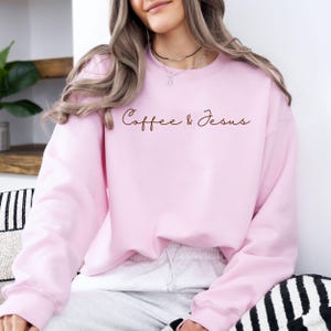 Puede incluir: Sudadera rosa claro con las palabras "Coffee & Jesus" en fuente marr&oacute;n. La sudadera de cuello redondo es de color liso. La persona que lleva la sudadera est&aacute; sentada.