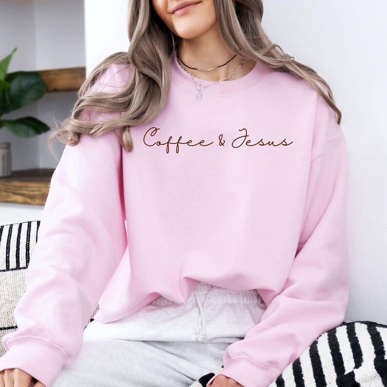 Puede incluir: Sudadera rosa claro con las palabras "Coffee & Jesus" en fuente marr&oacute;n. La sudadera de cuello redondo es de color liso. La persona que lleva la sudadera est&aacute; sentada.