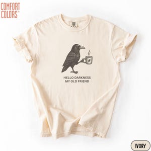 May include: An ivory short-sleeve t-shirt featuring a black graphic of a crow holding a steaming mug. Below the crow, the text reads HELLO DARKNESS MY OLD FRIEND. The shirt has a crew neckline and a relaxed fit, with a Comfort Colors brand label.