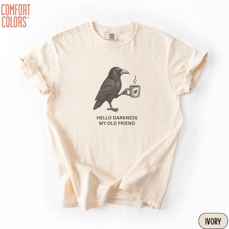 May include: An ivory short-sleeve t-shirt featuring a black graphic of a crow holding a steaming mug. Below the crow, the text reads HELLO DARKNESS MY OLD FRIEND. The shirt has a crew neckline and a relaxed fit, with a Comfort Colors brand label.