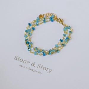 May include: A delicate gold bracelet with multiple strands of small, light blue and clear beads. The bracelet features a gold clasp and small gold heart charm. The words "Stone & Story handcrafted jewelry" are printed below the bracelet.