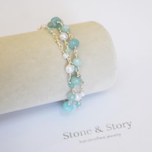 May include: A multi-strand bracelet featuring light blue and green gemstone beads interspersed with small white pearls on a thin gold-tone metal chain. The text Stone & Story handcrafted jewelry is printed in dark gray below the jewelry display.