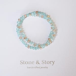 May include: A circular bracelet composed of faceted light blue and champagne-colored beads arranged in a repeating pattern. Below the bracelet, the text Stone & Story handcrafted jewelry is printed in a simple, dark gray serif font on a plain background.