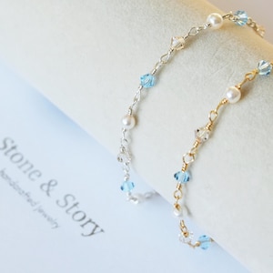 May include: Two delicate bracelets featuring alternating white pearls and light blue and clear faceted crystals on thin metallic chains. One chain is silver-toned and the other is gold-toned. The text Stone & Story handcrafted jewelry is visible.