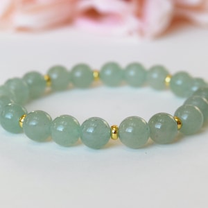 May include: A beaded bracelet featuring pale green, round gemstones. The beads are separated by small, gold-colored spacers. The bracelet is displayed on a white surface, with a blurred pink floral background.