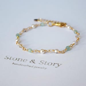 May include: Delicate gold-toned bracelet featuring small, round pearls, light green beads, and clear crystal accents. The bracelet is strung on a delicate gold chain and has a gold clasp. The words "Stone & Story handcrafted jewelry" are printed below the bracelet.