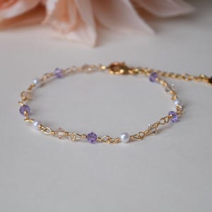 May include: Delicate gold-toned bracelet featuring small, clear, and purple beads, along with a few white pearls. The bracelet has an adjustable chain and a heart-shaped charm at the end. A beautiful piece of jewelry.