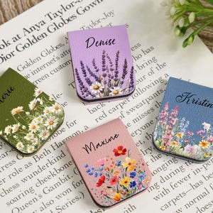 Puede incluir: Cuatro marcadores magnéticos personalizados con diseños florales. Cada marcador presenta un nombre: "Valerie" en verde, "Denise" en lavanda, "Maxine" en rosa y "Kristin" en azul. Los marcadores están sobre una página de libro.