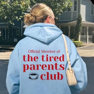 May include: A light blue hoodie with the text "Official Member of the tired parents club" in red. The hoodie features a coffee cup graphic and a small, off-white purse. The brand name "HELL BLAU" is printed on the left side.