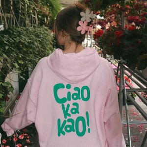 Puede incluir: Sudadera con capucha rosa claro con el texto "Ciao ka kao!" en verde en la espalda. La sudadera con capucha forma parte de una colección de diseño personalizado. La imagen también incluye un logotipo y las palabras "Light Pink/Back".