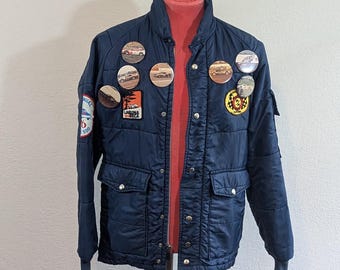 May include: A navy blue quilted jacket with multiple round and rectangular patches. The jacket has a red collar and is displayed on a metal stand. The jacket has four front pockets and a button-up closure.